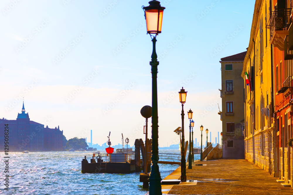 Sunlight bathes a Venetian promenade, where vintage lamp posts line the waterfront beside historic buildings. Tranquil canal reflects the golden hues of sunset, capturing timeless charm of Venice