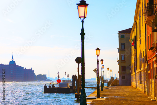 Obraz na płótnie Sunlight bathes a Venetian promenade, where vintage lamp posts line the waterfront beside historic buildings. Tranquil canal reflects the golden hues of sunset, capturing timeless charm of Venice w salonie