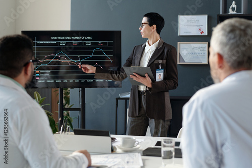 Side view portrait of mature female expert giving presentation to doctors during medical conference and pointing at statistics data on digital board copy space