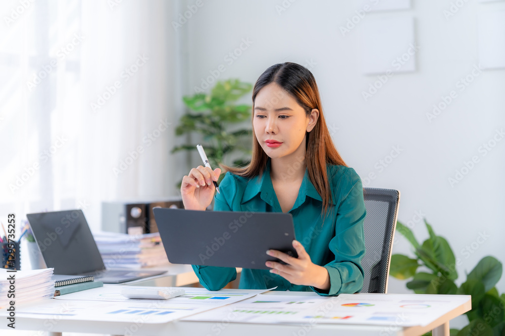 Focused Professional: An Asian woman meticulously reviews documents at her desk, surrounded by business essentials, the image portraying diligence and concentration. Focused Professional: An Asian woman meticulously reviews documents at her desk, surrounded by business essentials, the image portraying diligence and concentration.