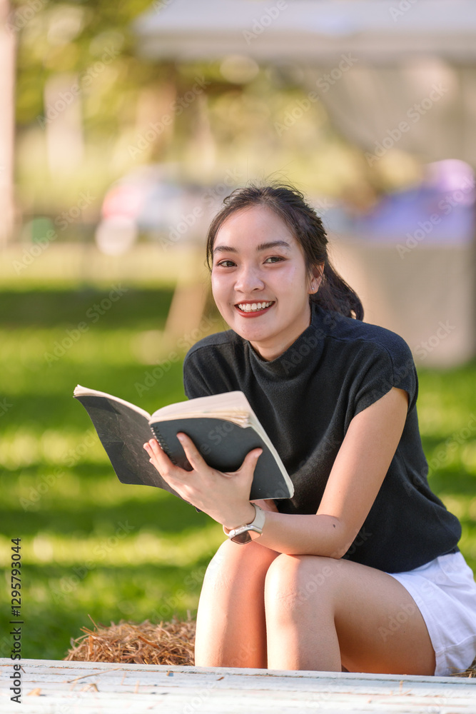 Fototapeta premium Young woman enjoys reading a book outdoors, sitting on a hay bale in a serene park environment, creating a tranquil scene of leisure and learning