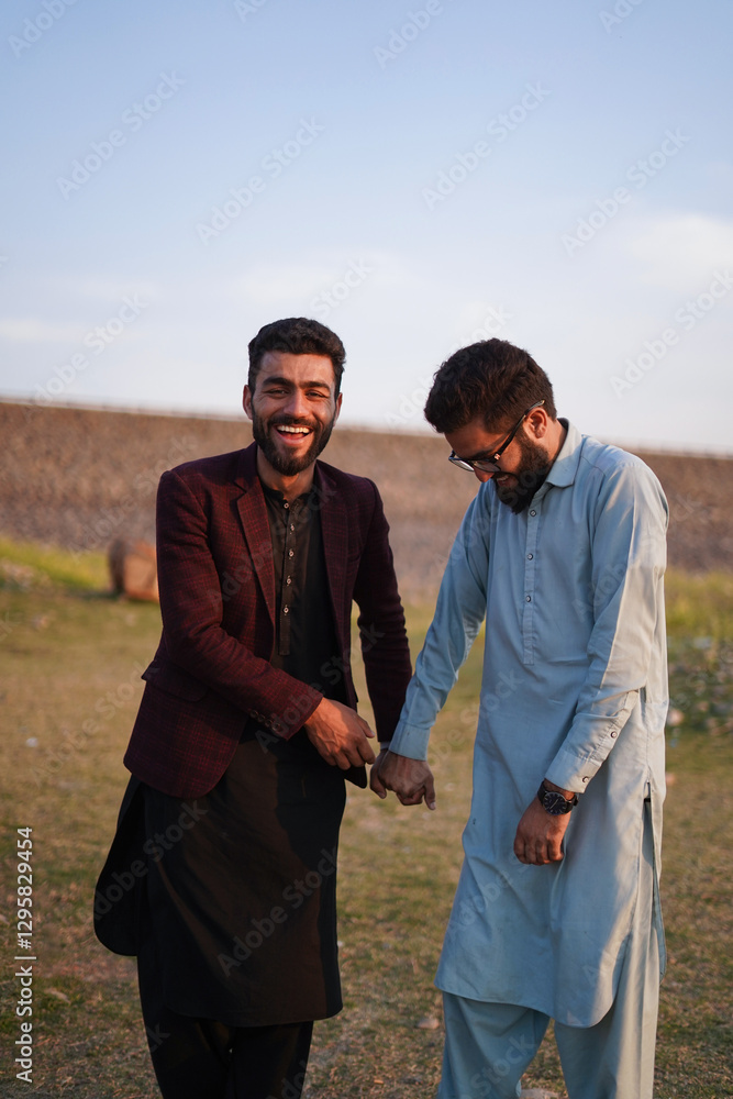 Two young South Asian men dressed in traditional and modern attire share a joyful moment, laughing together in an outdoor setting. Two young South Asian men dressed in traditional and modern attire share a joyful moment, laughing together in an outdoor setting.