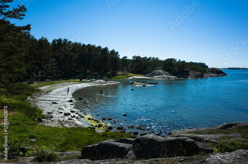 Small crescent-shaped beach on Rossö Island, Sweden. Rocky cliffs, shallow water, and a pine forest meet the blue sea under a clear summer sky. A serene coastal landscape