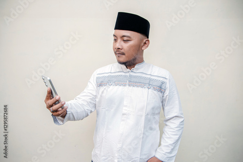 An Asian man wearing Muslim clothes and a peci makes a video call on Eid al-Fitr because he cannot go home to his hometown. close-up of Muslim man making video call isolated on white background photo