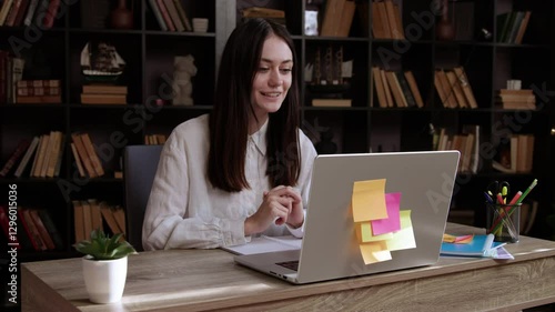 Young smiling brunette woman participates in an online interview and enthusiastically answers questions. Woman sits at an office table and talks in front of a computer explaining with her hands