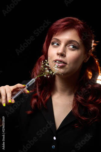 Obraz na płótnie Woman with red hair, holding delicate white flowers in syringe, posing elegantly against dark background, artistic contrast concept w salonie