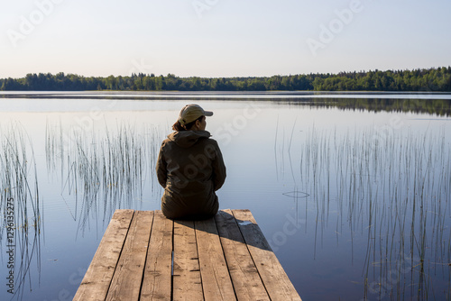 Fototapeta Naklejka Na Ścianę i Meble -  A woman sits on a wooden walkway on the shore of a beautiful lake and admires nature. Morning on a lake in the countryside. Traveling and hiking in nature. Outdoor recreation. Peace and quiet.