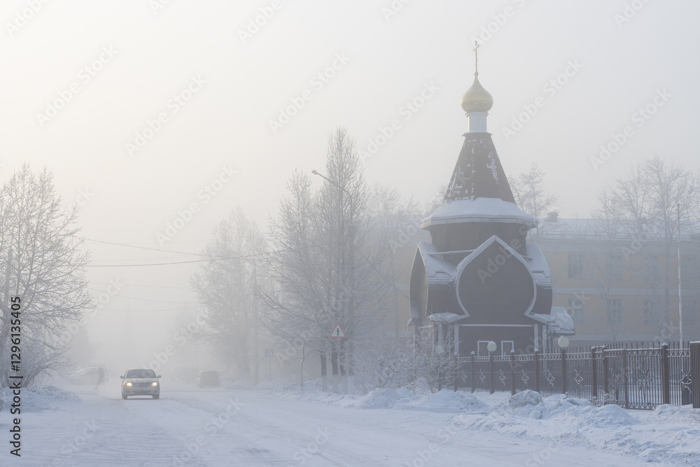 Fototapeta premium Winter landscape with snow-covered street and Orthodox church. Car is driving along the road. Very cold weather. Winter frost. Frosty fog. City of Kyzyl, Republic of Tyva (Tuva), Siberia, Russia.