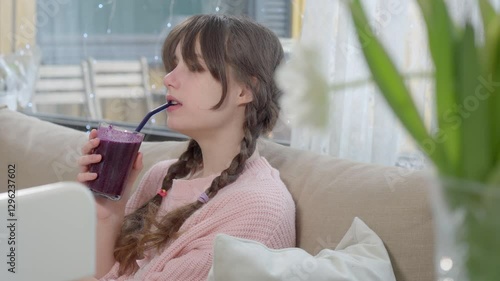A teenage girl in a pink knitted sweater is drinking a freshly blended berry smoothie from a glass. Healthy nutrition, homemade drinks, and a balanced diet for kids and teenagers
