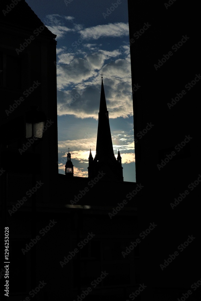 Obraz premium The tower is a tower with a high spire at the end of which a cross is seen among the buildings against the blue sky with gray clouds and bright sunlight.