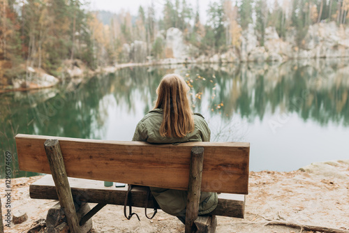 Lonely 30s blonde woman is sitting on the bench of a forest lake. Autumn landscape, calmness, tranquility, travel concept. A woman on the bank of a lake is enjoying the nature. High quality photo.