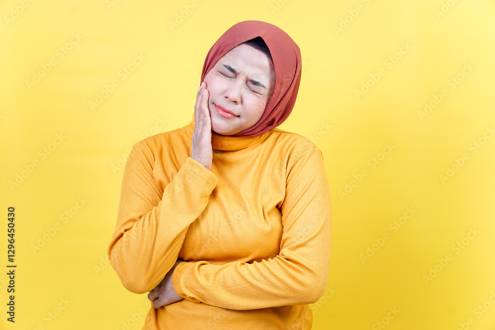 Fototapeta premium An asian woman in a hijab and yellow sweater is experiencing tooth pain, pressing her cheek with a pained expression.