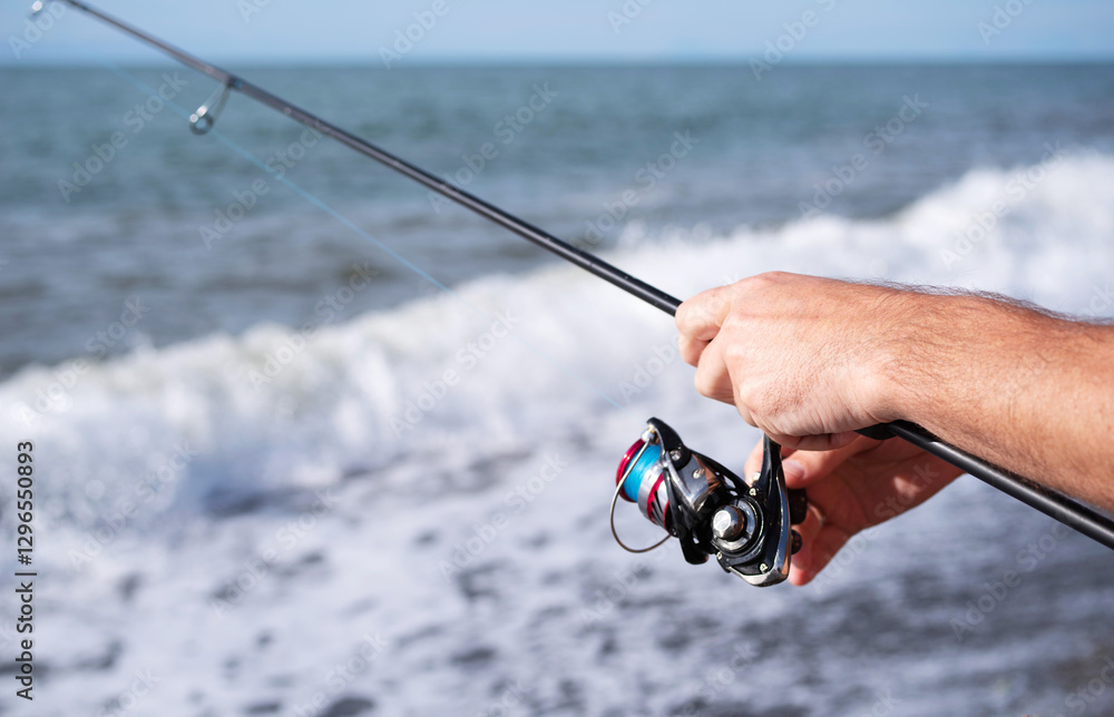 Spinning fishing. Close-up of male fisherman hands catching fish with spinning rod at sea on sunny hot day against blue sky background Spinning fishing. Close-up of male fisherman hands catching fish with spinning rod at sea on sunny hot day against blue sky background
