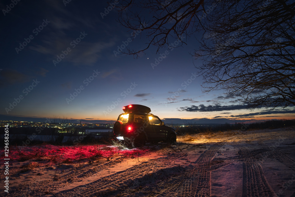 an old all-terrain vehicle stands on the top of the mount, In the background are the lights of the city, a late sunset on a winter evening. Glowing running lights and red stops. Stars in the sky