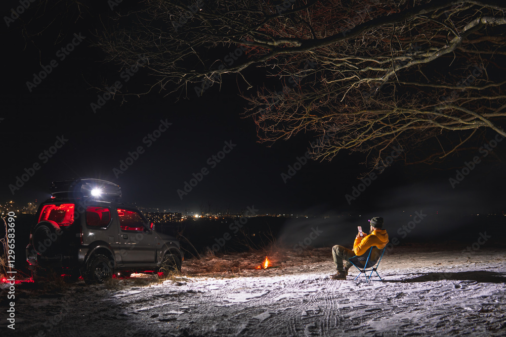 A man sits on a chair with a view of the lights of the night city. A bonfire burns with smoke. An off-road vehicle is parked on top. The concept of recreation and trips to nature. Use smartphone