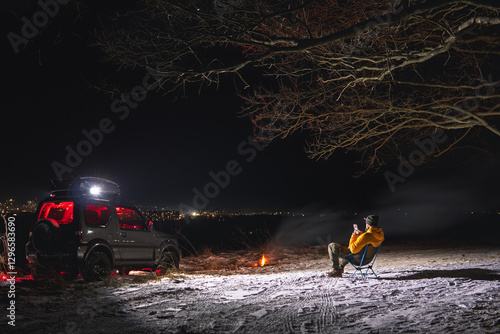 Obraz na płótnie A man sits on a chair with a view of the lights of the night city. A bonfire burns with smoke. An off-road vehicle is parked on top. The concept of recreation and trips to nature. Use smartphone w salonie