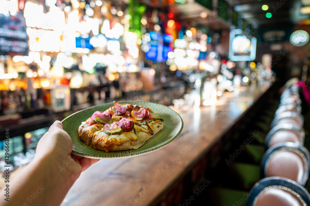 Fototapeta premium hand holds a plate of pizza in a vibrant bar setting, with a blurred background showcasing drinks and colorful lights, creating a lively atmosphere. 