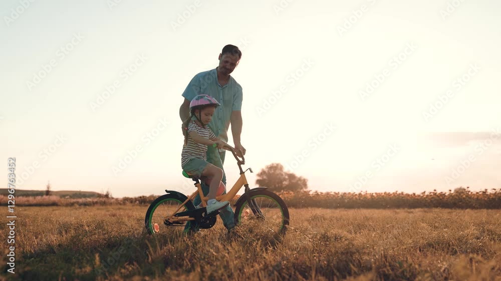 Attentive father supports little girl learning to ride bicycle in sunset field. Little girl in helmet trains bicycle riding with father in park. Happy father and girl plays with bicycle on holiday Attentive father supports little girl learning to ride bicycle in sunset field. Little girl in helmet trains bicycle riding with father in park. Happy father and girl plays with bicycle on holiday