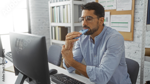 Картината върху платно Young hispanic man in office thinking with mobile against his mouth, sitting by a computer in a modern indoor workplace setting, reflecting professionalism and focus