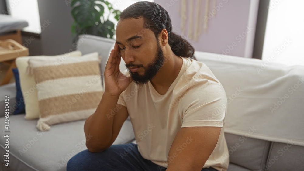 Young man with beard sitting in a living room looking thoughtful, surrounded by soft furnishings, creating a calm and introspective atmosphere in an indoor setting. Young man with beard sitting in a living room looking thoughtful, surrounded by soft furnishings, creating a calm and introspective atmosphere in an indoor setting.