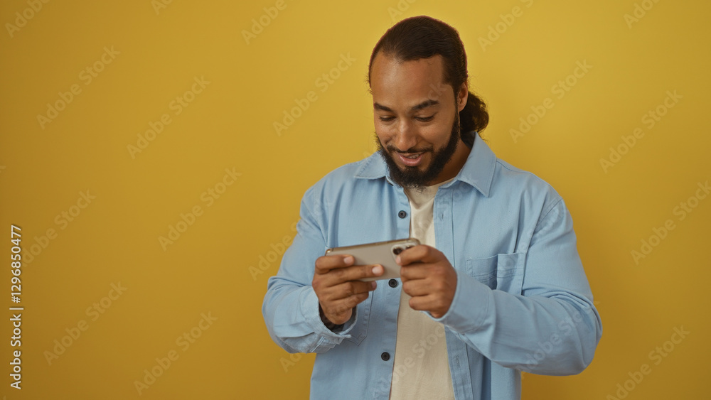 Man smiling at smartphone over yellow background, wearing blue shirt, beard visible, playing game or watching video, isolated in vibrant setting, adult enjoying digital content. Man smiling at smartphone over yellow background, wearing blue shirt, beard visible, playing game or watching video, isolated in vibrant setting, adult enjoying digital content.