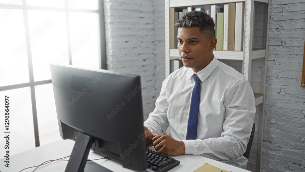 Handsome young hispanic man working in an office room typing on a computer with shelves and files in the background Handsome young hispanic man working in an office room typing on a computer with shelves and files in the background