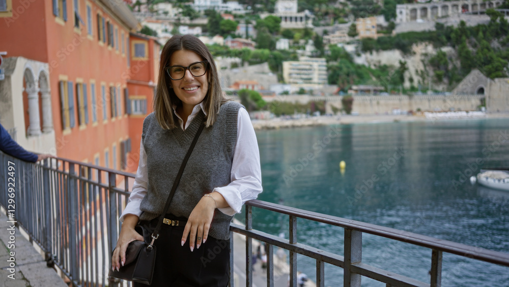 Obraz premium Hispanic woman smiling by the sea in villefranche france with scenic european backdrop wearing glasses and casual outfit highlighting travel and leisure vibes.