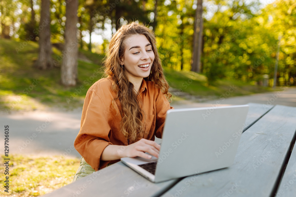 Cheerful woman sitting at a wooden table in the park using her laptop. Young female freelancer working in the park by the water. Leisure, business concept. Cheerful woman sitting at a wooden table in the park using her laptop. Young female freelancer working in the park by the water. Leisure, business concept.
