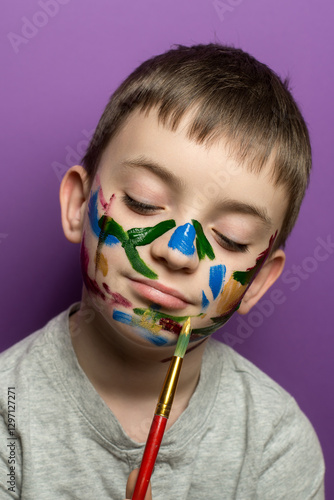 Wallpaper Mural Thoughtful young boy in a gray sweater with colorful face paint, holding a paintbrush against a purple background, perfect for childhood, creativity, and artistic expression themes in imagery. Torontodigital.ca