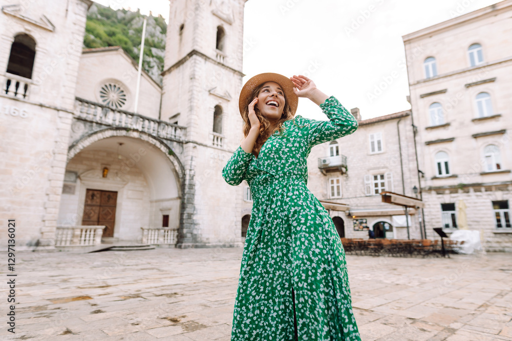 Obraz premium A cheerful woman in a green dress and a wide-brimmed hat is spinning in a city square. A young tourist enjoys a walk along European streets. Travel, vacation concept.