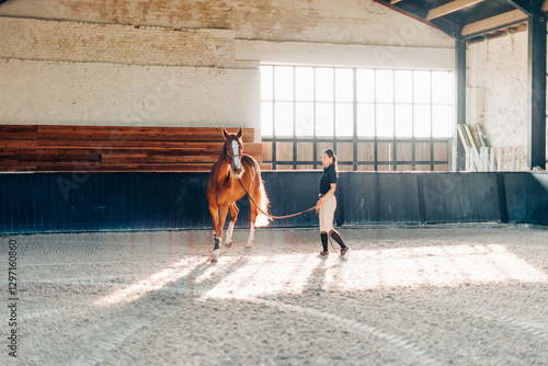 Female equestrian training a chestnut horse in an indoor riding arena with warm sunlight and rustic surroundings
