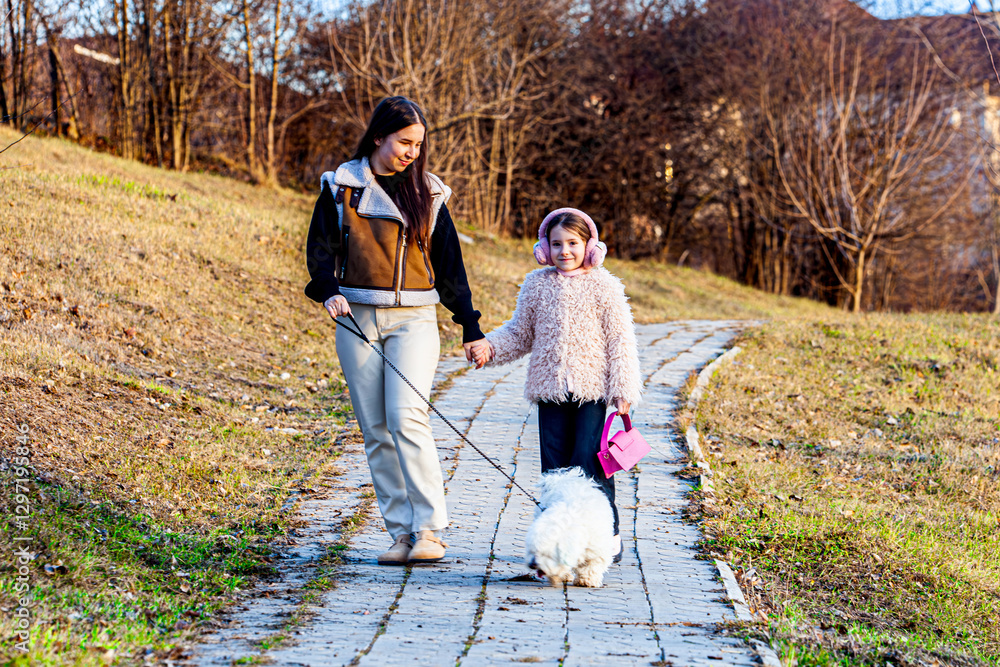 Fototapeta premium Two cute little girls were caught walking with a very fluffy white dog in a green park. The girls were walking with the dogs with enormous pleasure.