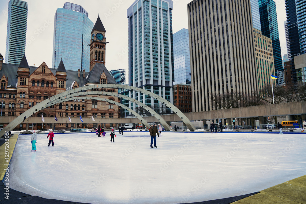 Fototapeta premium Toronto residents enjoy skating on the ice rink at the Nathan Phillips square,with the Old City Town Hall during winter in Toronto,Ontario,Canada 