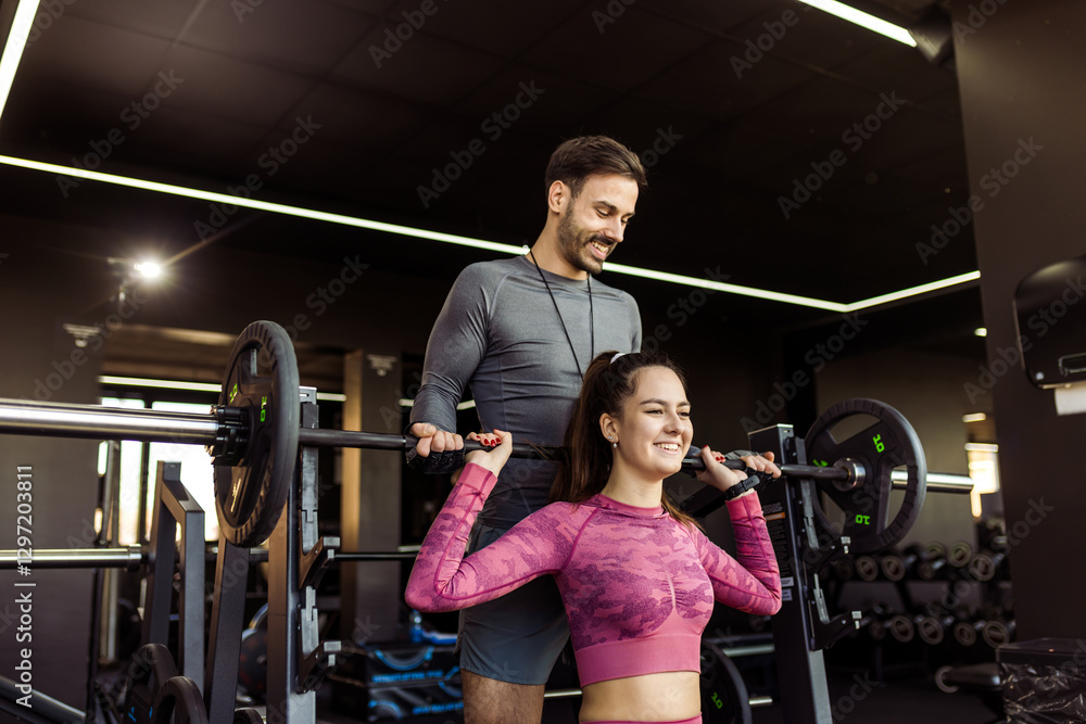 Fototapeta premium Young woman does shoulder press with weights in the gym while her personal trainer is spotting her.