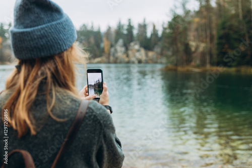 Person in hat is recording content with a smartphone near lake on wild nature background. Stylish 30s girl with phone walking outdoors. Travel, adventure. Concept of an active lifestyle.