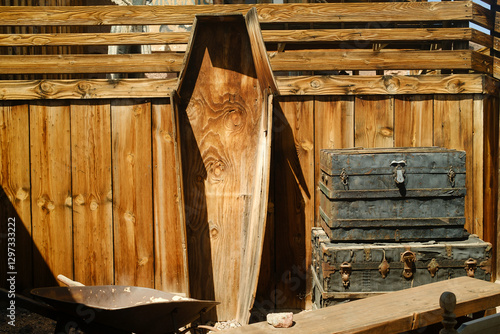 A weathered wooden coffin leans against a rustic fence, next to old metal trunks and a wheelbarrow