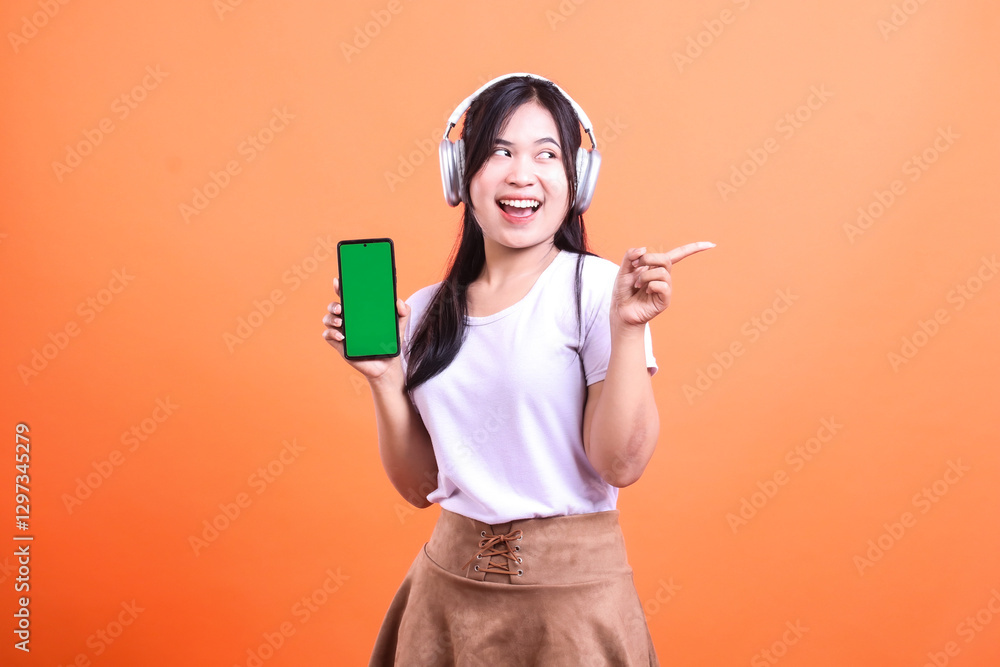 Smiling woman with headphones holding a smart phone device with green screen pointing to the side Smiling woman with headphones holding a smart phone device with green screen pointing to the side