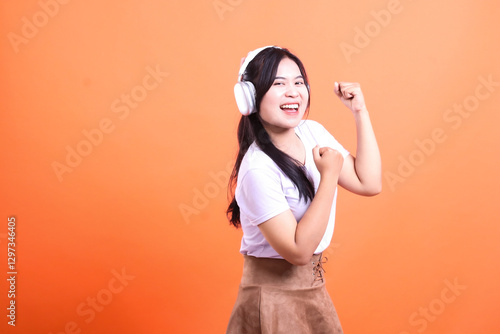 Woman listening music with white headphones and brown skirt posing against an orange background