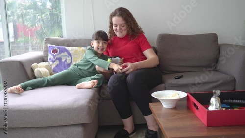A mother and daughter play video games together in the living room, trying to outsmart each other to win. they laugh and tease each other while enjoying their competitive moment.