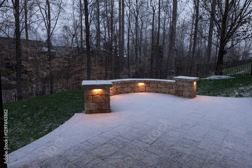 A peaceful backyard patio covered in fresh snow, featuring a curved stone wall with glowing lights. Black railings and bare trees enhance the serene winter ambiance at dusk.
