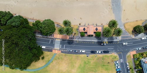 Fototapeta Naklejka Na Ścianę i Meble -  High-angle view of a beachside street scene. Cars parked along the road, pedestrians crosswalk, and a small building near the beach. Sunny day. SAINT HELIERS, AUCKLAND, NZ