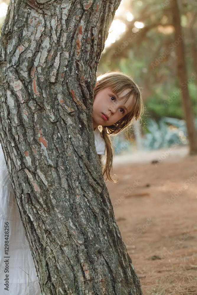 Child girl in white clothes peeking out from behind the tree in the park Child girl in white clothes peeking out from behind the tree in the park