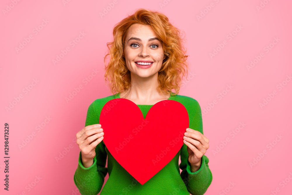 Bright cheerful woman holding a red heart symbol against a pink background, wearing a green sweater and showcasing a joyful gesture. Bright cheerful woman holding a red heart symbol against a pink background, wearing a green sweater and showcasing a joyful gesture.