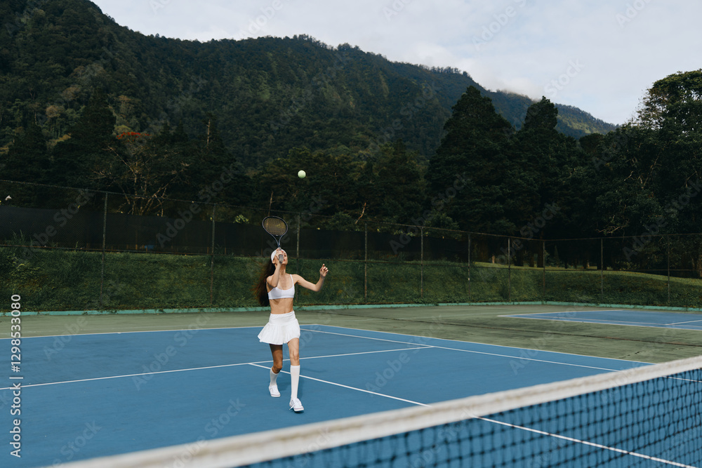 Obraz premium Female tennis player in white sportswear serving on outdoor blue court surrounded by greenery, showcasing determination and focus