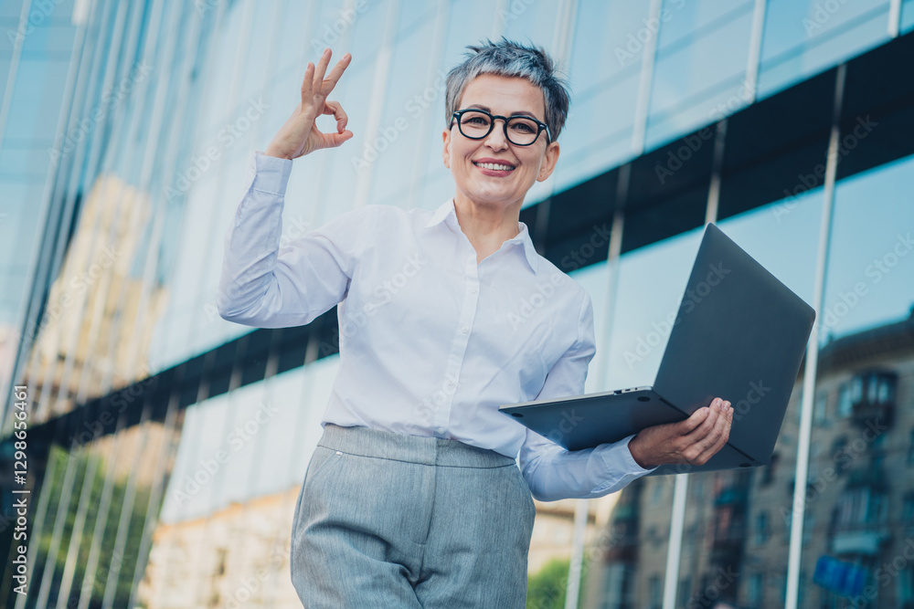 Confident middle-aged businesswoman with grey hair standing outdoors holding laptop and gesturing success in urban cityscape Confident middle-aged businesswoman with grey hair standing outdoors holding laptop and gesturing success in urban cityscape
