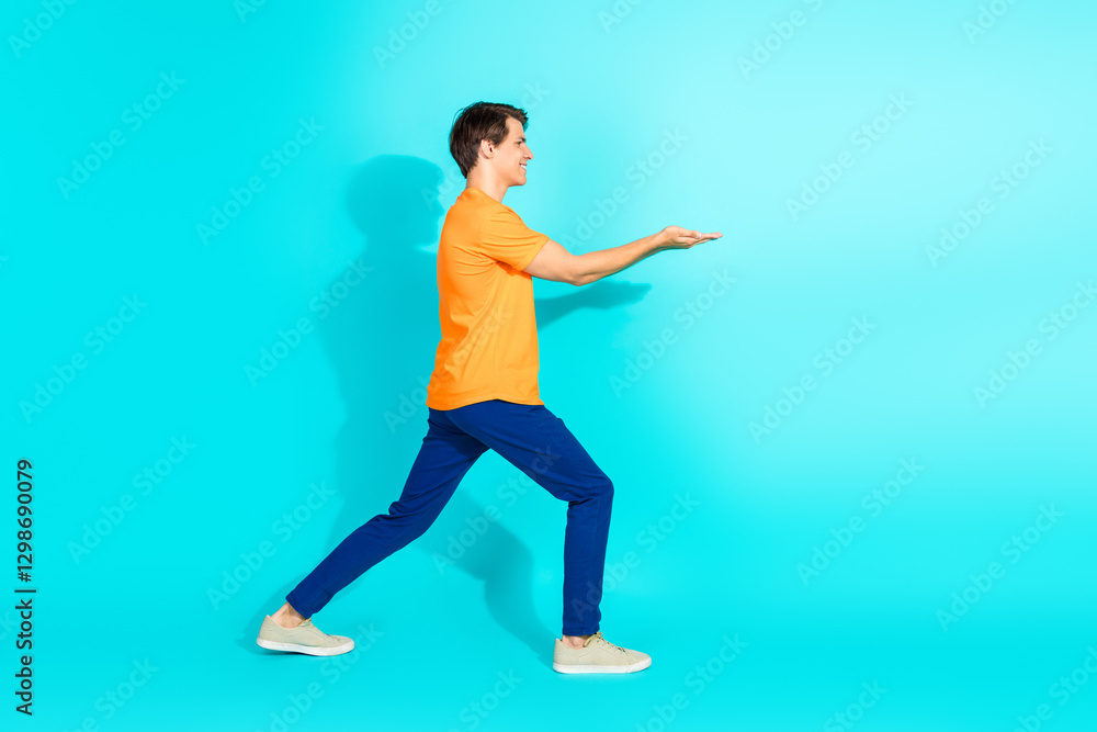 Obraz premium Energetic young man in an orange T-shirt and blue pants making a step with an open-hand gesture against a teal background