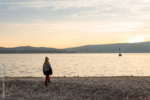 A girl in a jacket and red trousers stands on the shore of the Mediterranean Sea. The beach in the city of Tivat in Montenegro. Sunset, blue sky, early spring