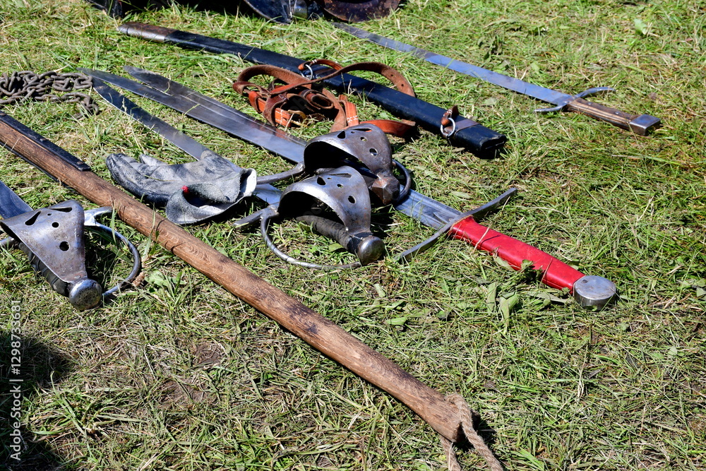 Naklejka premium A close up on a set of medieval weapons and accessories, including swords, hammers, pikes, targes, furs, bucklers and drums laying on the grass seen on a sunny summer day during a historic fair