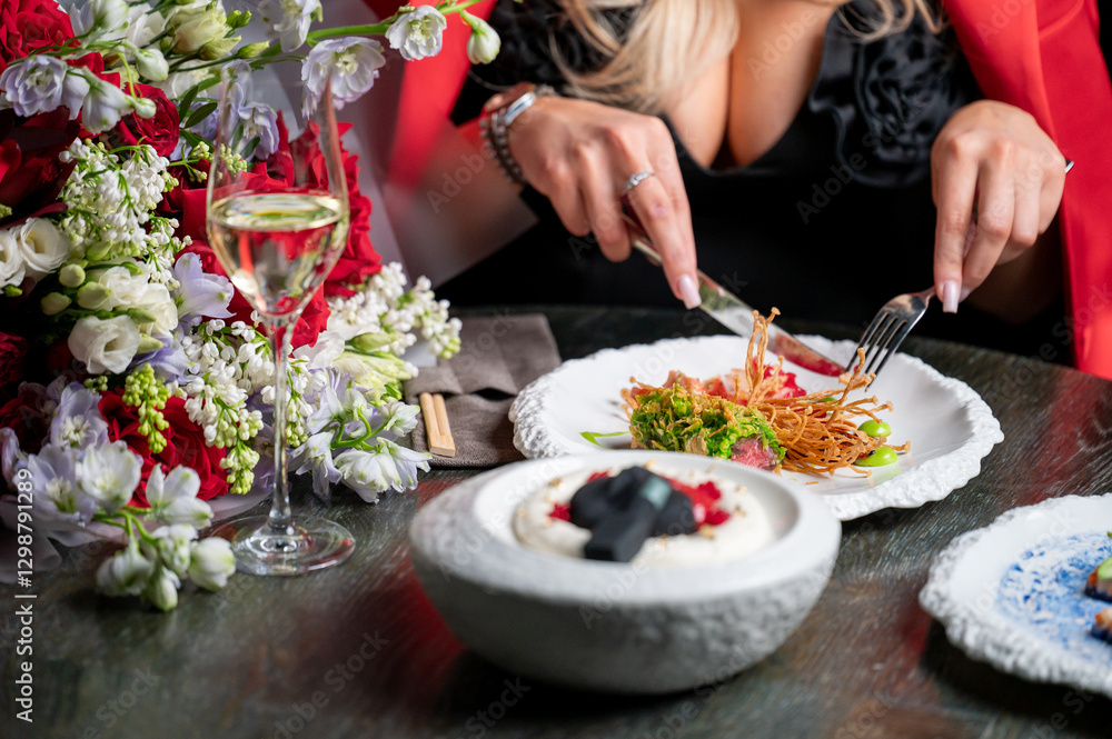 A close-up of a dining scene featuring a woman cutting her food with a fork and knife. Elegant dishes and flowers adorn the table, with a glass of champagne nearby, creating a luxurious atmosphere. A close-up of a dining scene featuring a woman cutting her food with a fork and knife. Elegant dishes and flowers adorn the table, with a glass of champagne nearby, creating a luxurious atmosphere.