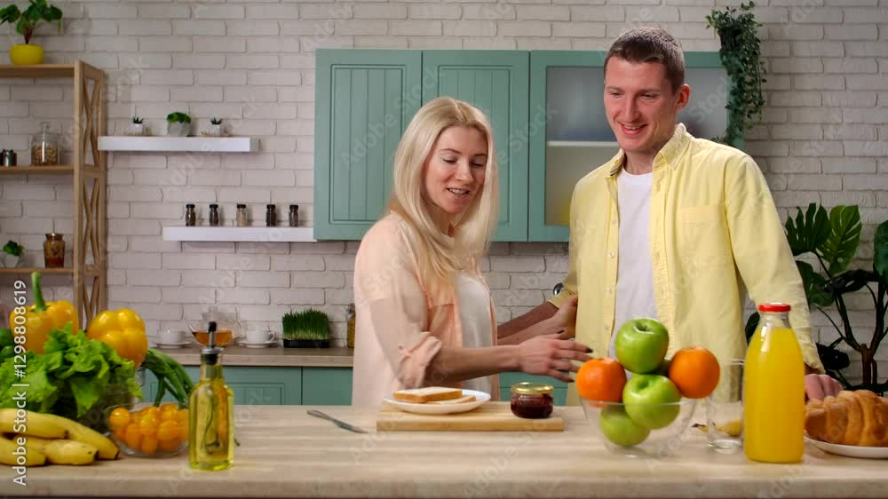 Couple of man and woman in the morning at home in the kitchen at table preparing breakfast, talking smiling, man helping woman make toasts with jam. Slow motion. Couple of man and woman in the morning at home in the kitchen at table preparing breakfast, talking smiling, man helping woman make toasts with jam. Slow motion.